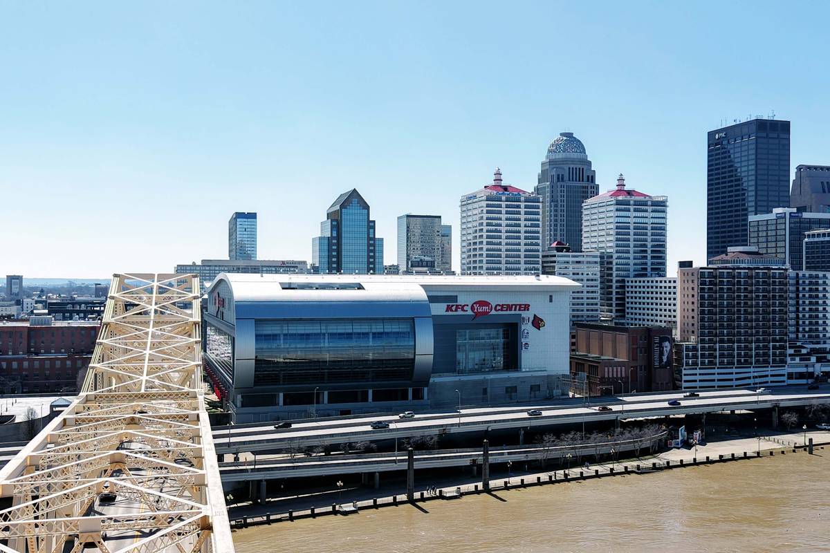 The Louisville skyline across the Ohio River, with a white steel truss bridge in the foreground and a waterfront arena at the base of the downtown towers.